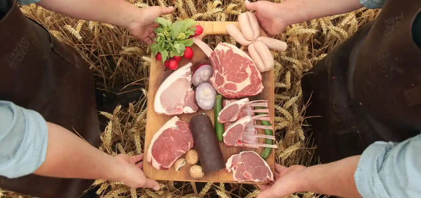 Two members of the Webster family holding a wooden board laden with award-winning cuts of beef, pork, lamb and homegrown vegetables, standing in a field of golden Lancashire wheat